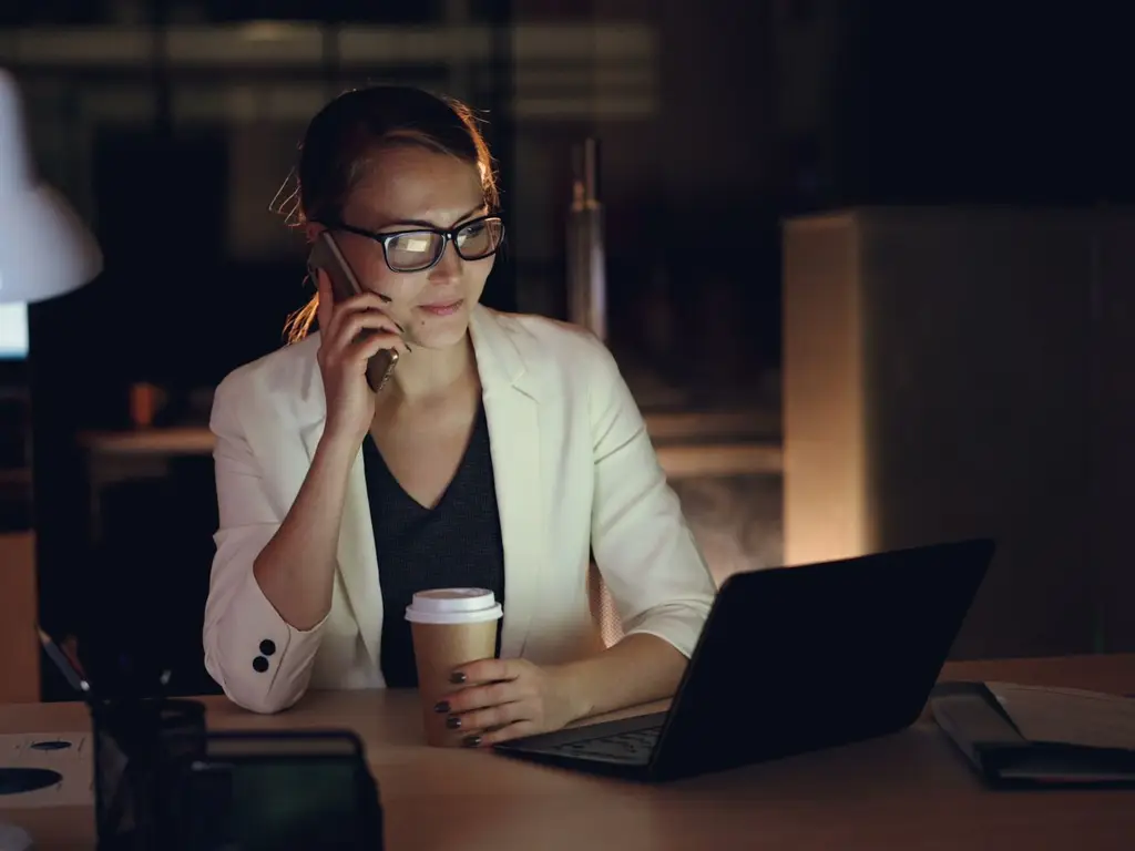 Woman in glasses working late at night on laptop, holding coffee, making phone call.