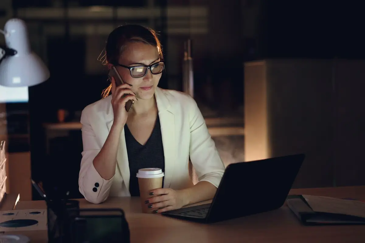 Woman in glasses working late at night on laptop, holding coffee, making phone call.